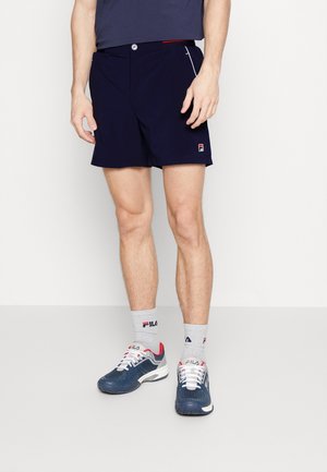 Mężczyzna w granatowych sportowych szortach, szarych skarpetkach FILA oraz granatowych butach sportowych FILA stoi na jednolitej jasnej podłodze na białym tle.