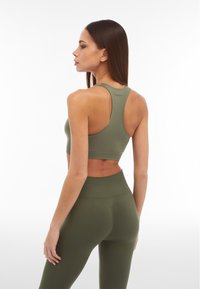 Donna in reggiseno sportivo verde e leggings sta rivolta di spalle, mostrando il design della parte posteriore dell'abbigliamento sportivo su uno sfondo bianco.