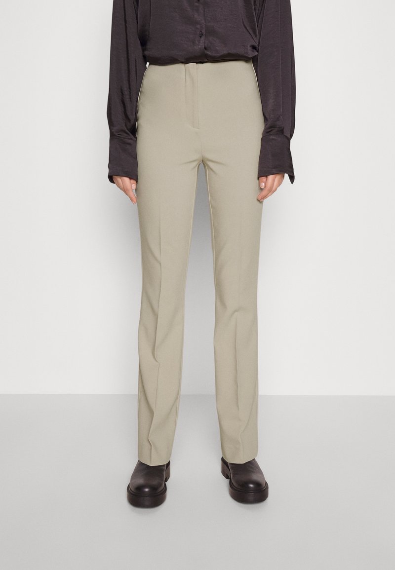 Personne portant un pantalon beige droit, une chemise noire à boutons avec de longues manches et des chaussures noires épaisses, debout devant un fond blanc.