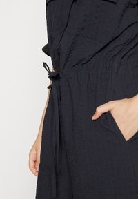 Gros plan d'une personne portant une robe noire texturée avec une ceinture à la taille, montrant une main dans une poche latérale sur fond blanc.