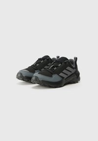 Schwarze und graue Sportschuhe mit einem strukturierten Obermaterial, verstärkter Zehenkappe und einer robusten Außensohle, die an den Seiten drei Streifen zeigen.