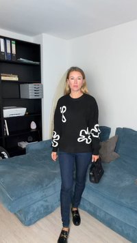 Črn pleten pulover z belimi valovitimi poudarki, kombiniran z temno modrimi kavbojkami in sijočimi črnimi loaferji. Sedi na teksturirani modri sedežni garnituri.