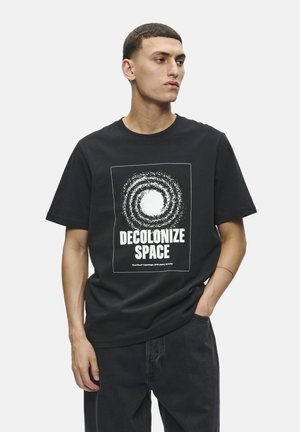 Camiseta de algodón negra con un estampado gráfico blanco que presenta el texto "Decolonizar el Espacio" y un diseño en espiral. Manga corta y corte relajado.