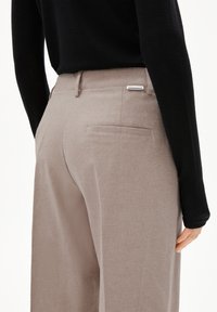 Relaxed-fit beige broeken gemaakt van een gestructureerde stof, met een achterzak en een labeldetail. Het model draagt een zwart langemouwen shirt.