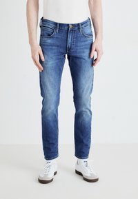 Jeans blu slim-fit realizzati in denim, con tasche anteriori e chiusura con zip. Il colore è un lavaggio chiaro a medio con lievi sbiaditure.