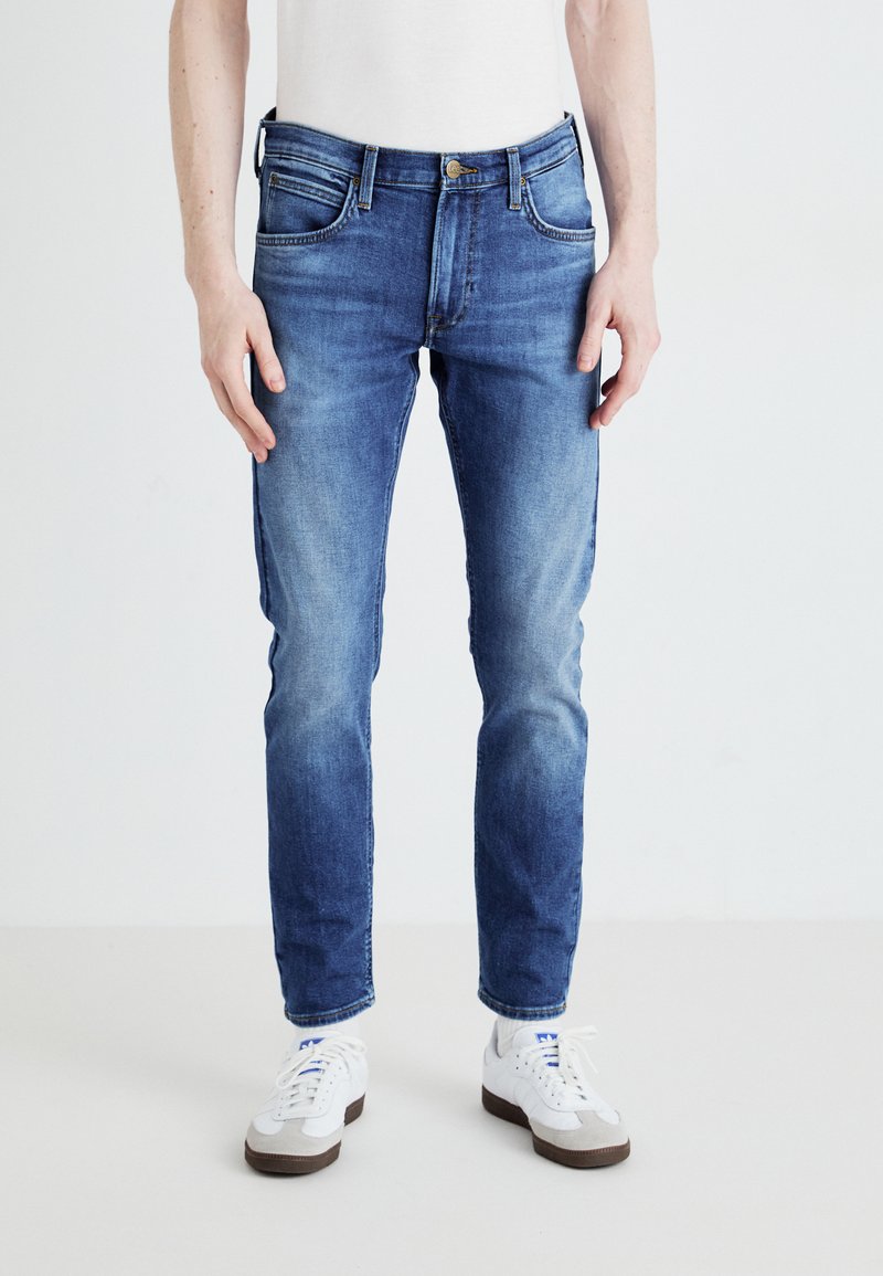 Jeans blu slim-fit realizzati in denim, con tasche anteriori e chiusura con zip. Il colore è un lavaggio chiaro a medio con lievi sbiaditure.