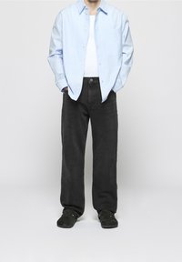 Camisa azul claro abotonada sobre una camiseta blanca sin mangas, combinada con vaqueros negros de corte amplio y calzado oscuro, creando un conjunto informal.