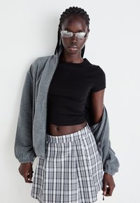Graue Zip-Jacke über einem schwarzen, gekürzten T-Shirt, kombiniert mit einem karierten Minirock in schwarz und weiß. Silberne Brille vervollständigt das Outfit.