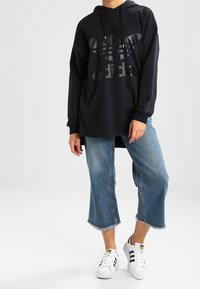 Svart huhoodtröja med präglad Adidas-logotyp, kombinerad med ljusblå denimculottes med fransade kanter och klassiska vita sneakers.