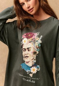 T-shirt oversize vert olive avec un portrait coloré imprimé de Frida Kahlo, agrémenté de fleurs et de la citation : "Je peins des fleurs pour qu'elles ne meurent pas."