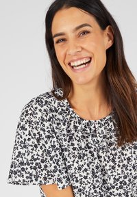 Witte blouse met zwart bloemmotief, korte fladdermouwen en een ronde halslijn. Stoffen lijkt lichtgewicht en gestructureerd.