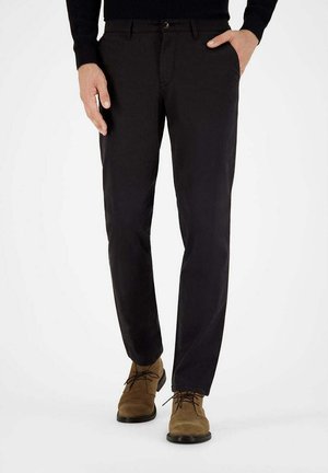 Mann trägt schwarze gerade geschnittene Hose und braune Wildleder-Schnürschuhe, steht mit einer Hand in der Tasche.