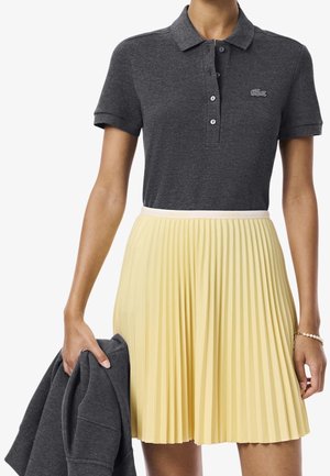 Kvinde iført mørkegrå polo og lysegult plisseret skørt, der holder en foldet mørkegrå jakke i venstre hånd.