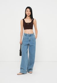 Brun cropped tank topp med logo, kombinert med lyseblå vide jeans. Modellen holder en liten svart veske, og står mot en hvit bakgrunn.