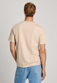 Camiseta beige de manga corta con un escote redondeado, hecha de una tela suave. La parte trasera tiene un diseño sencillo con un dobladillo recto y sin logotipos visibles.