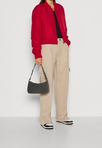 Rode bomberjack, zwart topje, beige broek met wijde pijpen, grijze suède handtas en zwart-witte sneakers. Zachte texturen, casual ontwerp.