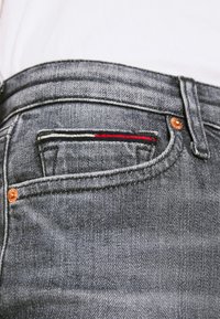 Jeans de mezclilla gris con un bolsillo delantero que presenta acentos de costura en rojo, blanco y azul, y detalles de herrajes de cobre.