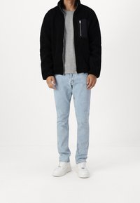 Schwarze Fleecejacke mit hohem Kragen und Reißverschluss-Brusttasche, über einem grauen T-Shirt getragen, kombiniert mit hellblauen Jeans und weißen Turnschuhen.