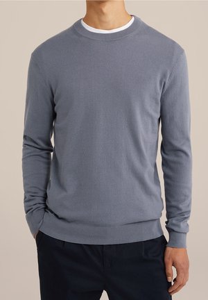 Homme portant un pull bleu clair à manches longues et col rond par-dessus une chemise blanche, avec un pantalon foncé et les mains dans les poches, sur un fond neutre.