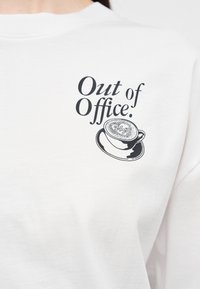 Λευκό πουκάμισο με μαύρο κείμενο "Εκτός Γραφείου." πάνω από μια εικόνα καφετιέρας με αφρώδες επάνω μέρος και τη λέξη "surf" στον αφρό.