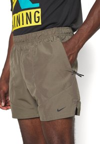Gros plan d'une personne portant un short de sport Nike vert olive avec taille élastique et main dans la poche latérale, associé à un t-shirt graphique noir et jaune.