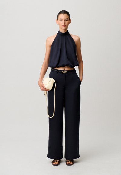 Donna con top nero senza maniche a collo alto e pantaloni neri a gamba larga e vita alta, che tiene una piccola borsa beige a tracolla, in piedi contro uno sfondo semplice.