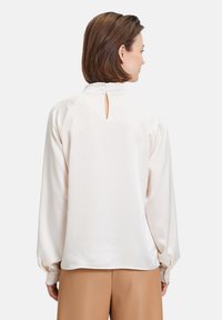 Blusa color crema con maniche lunghe e voluminosi dettagli a palloncino e apertura a chiave sul retro. Tessuto liscio con una vestibilità rilassata.