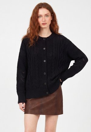 Donna con capelli rossi che indossa un cardigan nero strutturato con bottoni e una gonna di pelle marrone su uno sfondo chiaro e uniforme.