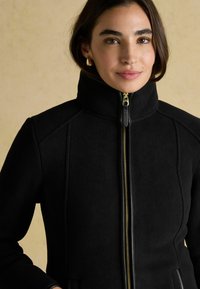 Žena s tmavými vlasmi, oblečená v zlatých náušniciach a čiernej bundy na zips s vysokým golierom, stojaca pred béžovým pozadím.