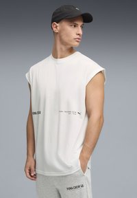 T-shirt blanc sans manches à col rond, avec un texte noir et un petit logo. Associé à un pantalon de survêtement gris, affichant un style sportif décontracté.