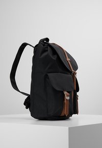 Zwarte stoffen rugzak met bruine leren accenten, een klepsluiting en een voorvak. Voorzien van verstelbare riemen en een zijrits.