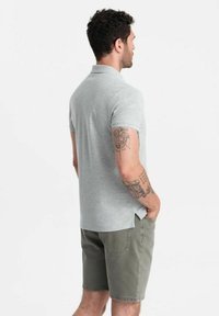 Grå kortärmad polo med platt krage, sidslitningar på nederkanten, buren med olivgröna shorts; har tatueringar på armen.