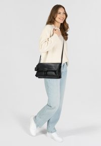 Borsa a tracolla nera in pelle con patta pieghevole, tasca con zip e tracolla regolabile, mostrata accanto a jeans blu chiaro e un maglione color crema.