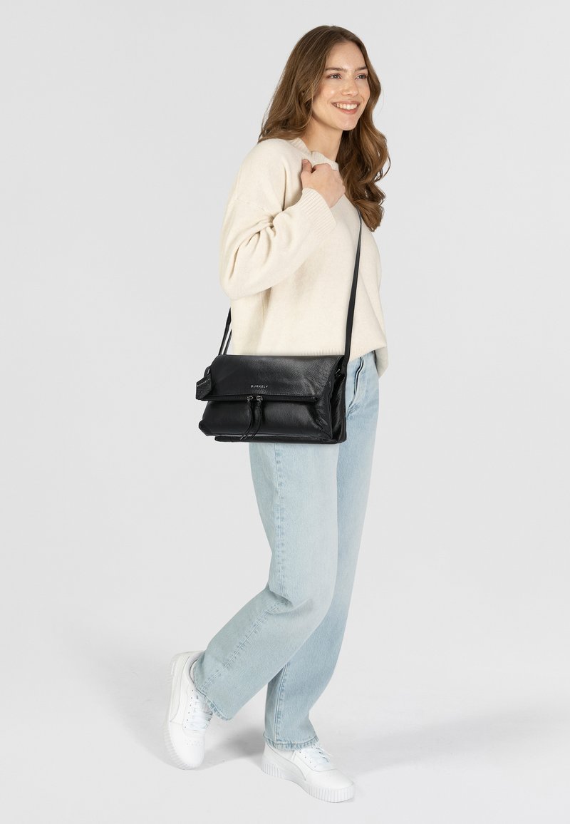Borsa a tracolla nera in pelle con patta pieghevole, tasca con zip e tracolla regolabile, mostrata accanto a jeans blu chiaro e un maglione color crema.