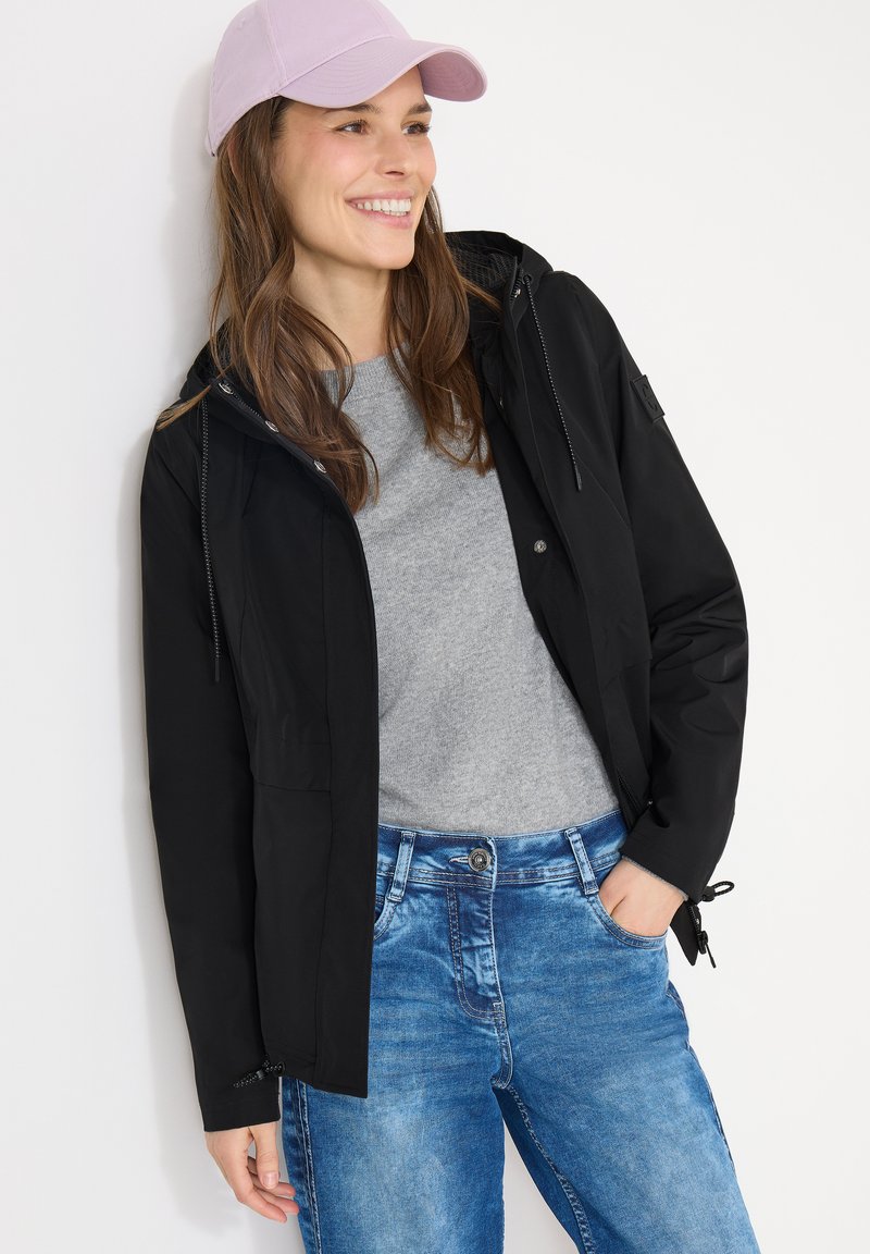 Femme souriante portant une casquette rose, une veste noire, un t-shirt gris et un jean bleu, une main dans la poche, sur un fond blanc.