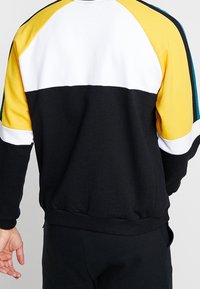 Sudadera negra con diseño en bloques de color amarillo, blanco y verde azulado. Presenta un cuello y puños acanalados, y un corte relajado.