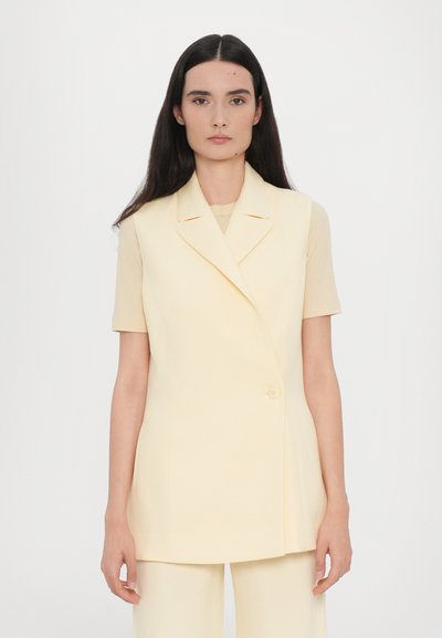 Blazer sans manches jaune avec un col cranté, fermeture à un bouton et coupe ajustée, associé à un t-shirt beige clair.