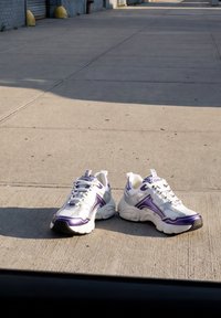 Scarpe da ginnastica bianche e viola posate su un marciapiede di cemento soleggiato vicino a un edificio con porte grigie chiuse.