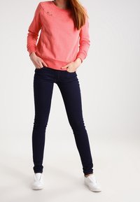 Felpa color corallo con scollatura rotonda e bottoni decorativi, abbinata a jeans skinny blu scuro e scarpe slip-on bianche.