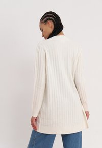 Long, ribbed cardigan in soft, light cream knit with side slits, fitted sleeves, and a straight hem. Worn over loose blue jeans.