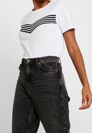 Persoon draagt een wit T-shirt met zwarte horizontale strepen en een hooggetailleerde vervaagde zwarte spijkerbroek, arm ontspannen langs het lichaam.