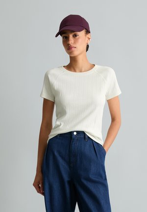 Frau mit einer bordeauxroten Kappe, weißem kurzärmligem Rippstrickshirt und hoch taillierter, weiter dunkelblauer Jeans mit einer Hand in der Tasche.