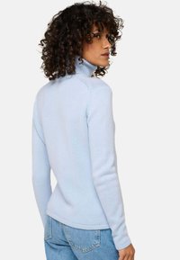 Femme aux cheveux bouclés portant un pull col roulé bleu clair et un jean bleu, se retournant sur son épaule devant un fond uni.