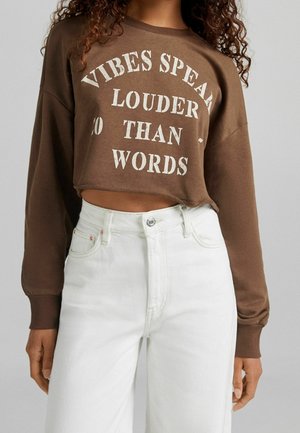Felpa corta marrone con testo bianco "LE EMOZIONI PARLANO PIÙ FORTE DELLE PAROLE" e maniche lunghe, abbinata a jeans in denim bianchi a vita alta.