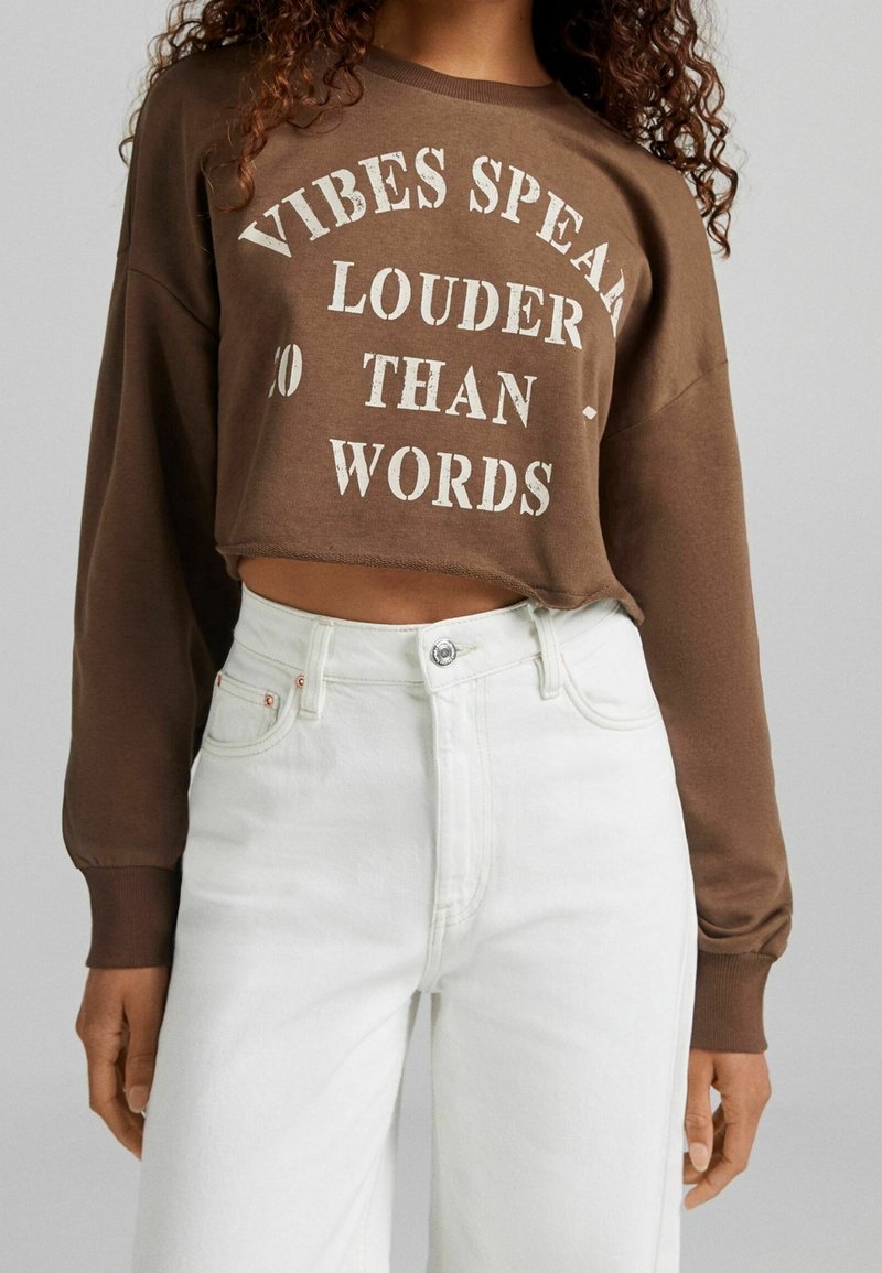 Felpa corta marrone con testo bianco "LE EMOZIONI PARLANO PIÙ FORTE DELLE PAROLE" e maniche lunghe, abbinata a jeans in denim bianchi a vita alta.