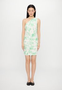 Robe asymétrique à une épaule en blanc avec un motif floral vert, design ajusté, texture froncée, longueur genou, associée à des chaussures plates noires.