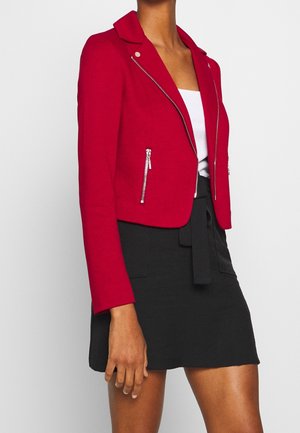 Femme portant un blazer rouge ajusté avec des détails de fermeture éclair argentée sur un top blanc et une jupe noire taille haute avec une ceinture nouée devant.