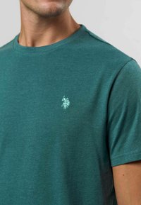 Mėlynos spalvos trumpomis rankovėmis marškinėliai, pagaminti iš medvilnės, su subtiliu logotipo siuvinėjimu kairėje krūtinės pusėje ir klasikiniu apvaliu iškirpimu.