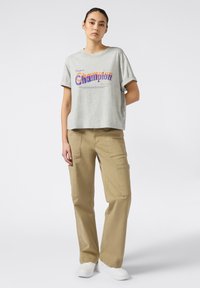 Camiseta gris corta con el logo "Champion" en púrpura y naranja, combinada con pantalones cargo color beige con bolsillos laterales, usados con zapatillas blancas.