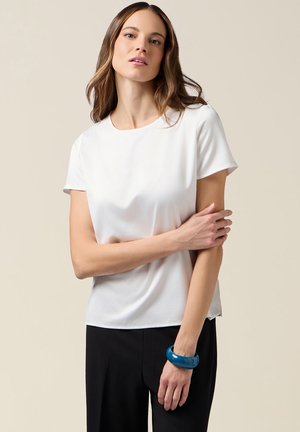 Top bianco a maniche corte in tessuto liscio, scollo rotondo, abbinato a pantaloni neri. La donna indossa un braccialetto blu al polso.